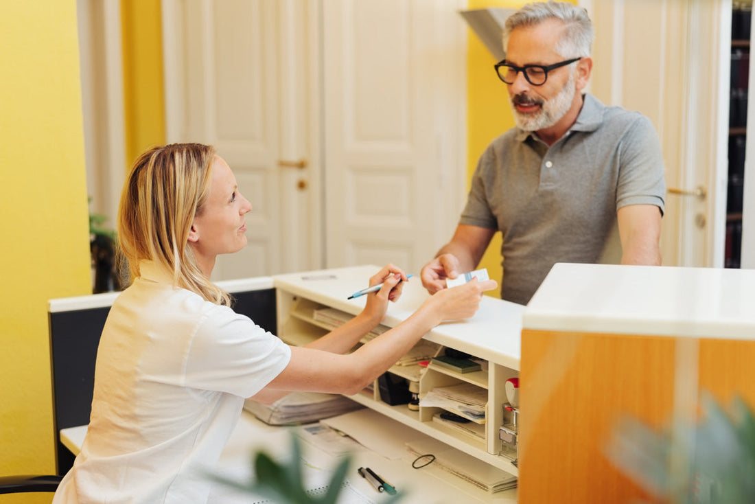 Friendly female receptionist handing paperwork to an older male patient at a clinic front desk, illustrating a welcoming healthcare office environment.