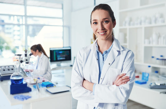 Smiling female scientist in a white lab coat standing confidently in a modern laboratory with arms crossed, while another researcher works with a microscope in the background.
